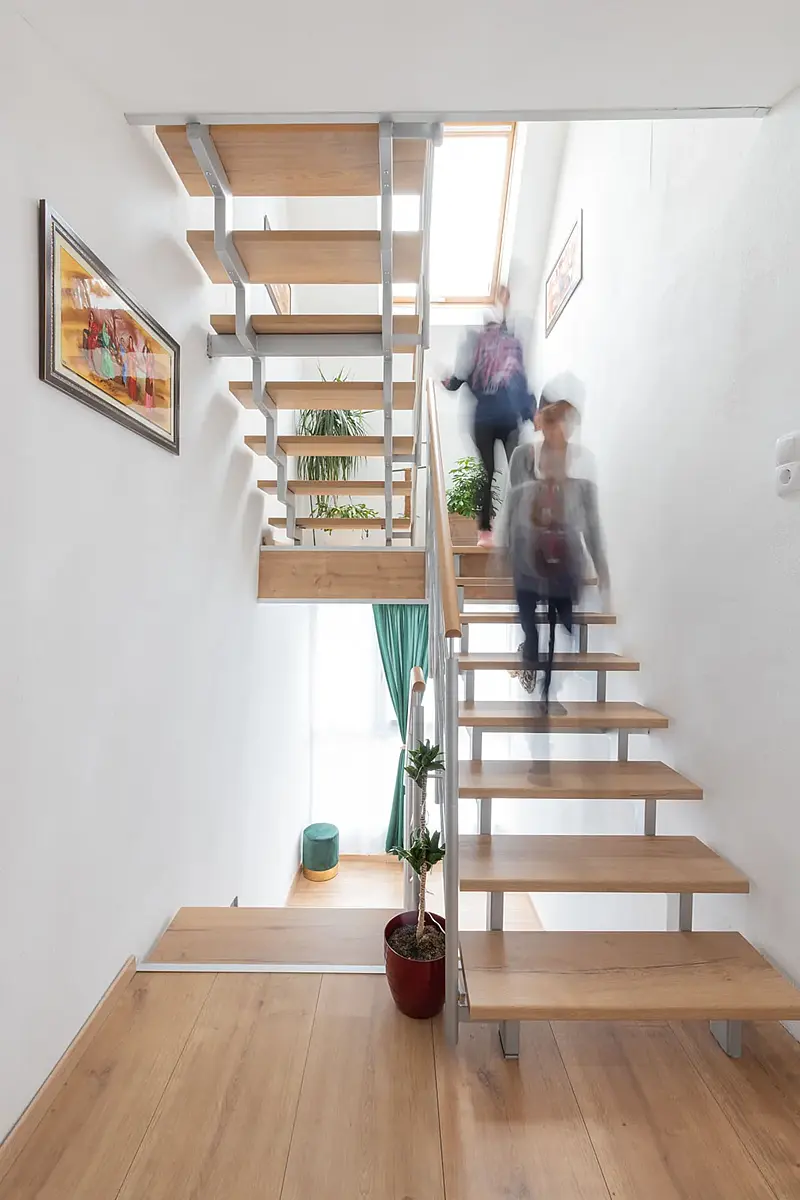 Blick ins Treppenhaus mit freitragender Wildeiche-Treppe; zwei Personen gehen als Bewegungsunschärfe die Stufen hinauf, an der Wand ein gerahmtes Bild.