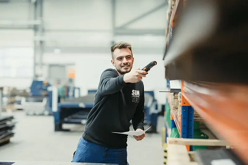 Lagermitarbeiter im Firmen-Hoodie scannt mit einem Handgerät Ware im Regal, im Hintergrund die weite, hell ausgeleuchtete Fertigungshalle.