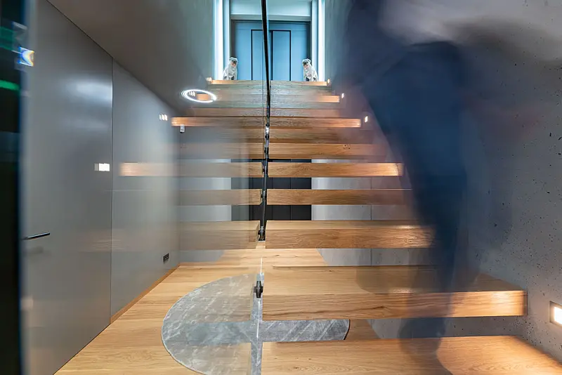 Kragarmtreppe aus Eiche an einer Sichtbetonwand, Ganzglasgeländer mit schwarzem Handlauf; rechts steigt eine Person als Bewegungsunschärfe hinauf, im Eichenboden ein runder Marmoreinsatz.