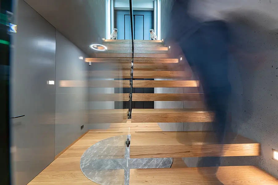 Kragarmtreppe aus Eiche an einer Sichtbetonwand, Ganzglasgeländer mit schwarzem Handlauf; rechts steigt eine Person als Bewegungsunschärfe hinauf, im Eichenboden ein runder Marmoreinsatz.