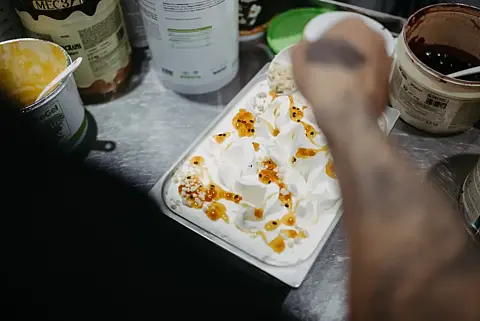 In einer Eismanufaktur zieht eine Hand Karamellsauce über eine frisch abgefüllte Wanne Vanilleeis, ringsum Zutatenbehälter auf dem Edelstahltisch.