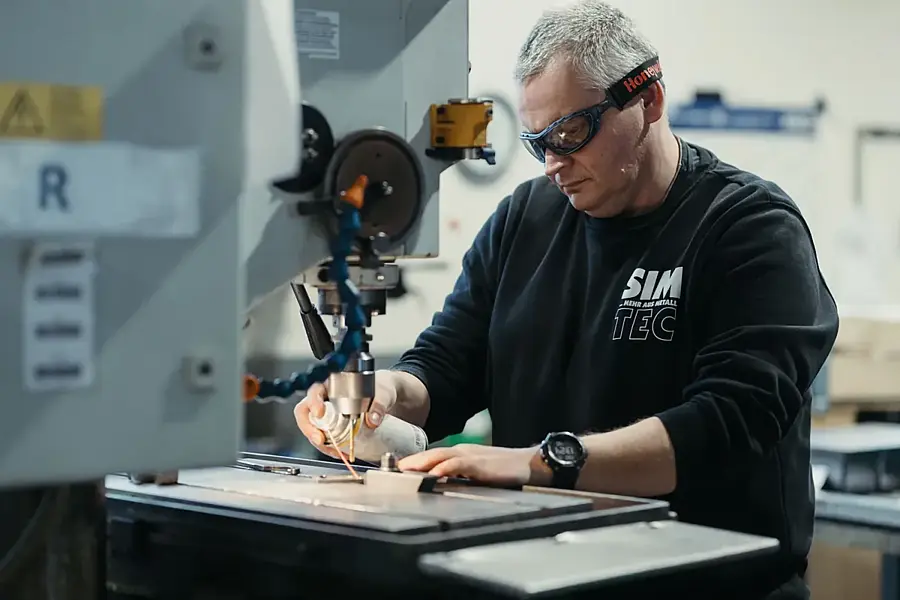 Metallbauer mit Schutzbrille und Gehörschutz führt an einer Ständerbohrmaschine ein Werkstück, Kühlmittel läuft über die Bohrstelle.