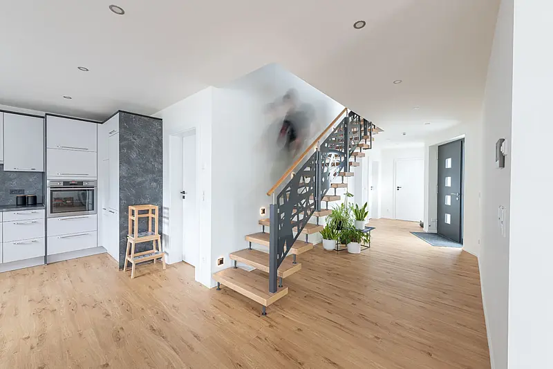 Offener Wohnbereich mit Eichendielen: links eine helle Küchenzeile, mittig eine Treppe mit Holzstufen und anthrazitfarbener Wange, an der jemand als Bewegungsunschärfe hinaufgeht.