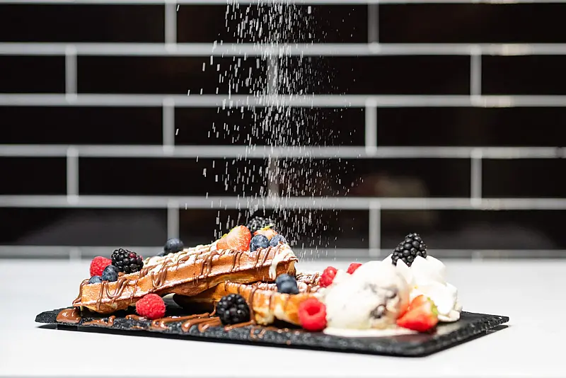 Puderzucker rieselt auf eine Waffelplatte mit Schokoladensauce, frischen Beeren und einer Kugel Eis, dahinter eine dunkle Fliesenwand.