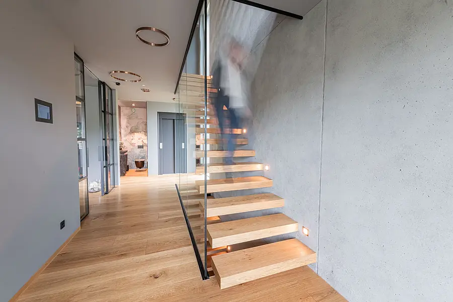 Heller Flur mit Eichendielen und Sichtbetonwand; rechts führt eine Kragarmtreppe mit Ganzglasgeländer nach oben, auf der jemand als Bewegungsunschärfe hinaufsteigt.