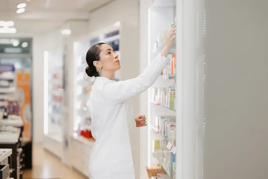 Apothekerin im weißen Kittel greift in der Offizin nach einer Packung im oberen Regalfach, ringsum hell beleuchtete Sichtwahl-Regale.