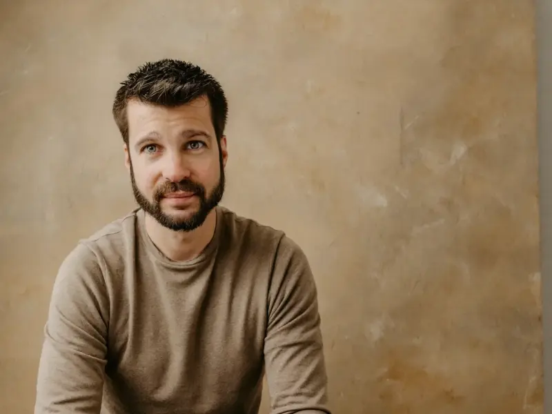 Armin Barth sitzt auf einem Hocker vor einem sandfarbenen Studiohintergrund, sandfarbener Feinstrick, Hände locker auf den Knien, ruhiger Blick in die Kamera.