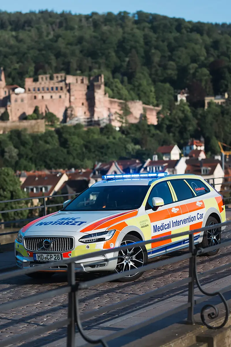 Notarztfahrzeug auf einer gepflasterten Brücke mit schmiedeeisernem Geländer, dahinter über den Dächern der Altstadt das Heidelberger Schloss.