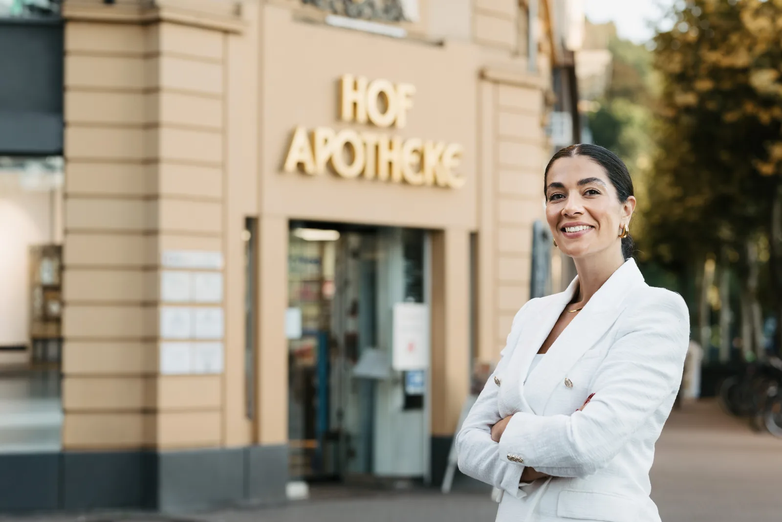 Apothekerin im weißen Blazer steht mit verschränkten Armen lächelnd vor ihrer Apotheke, an der Fassade hinter ihr der goldene Schriftzug.