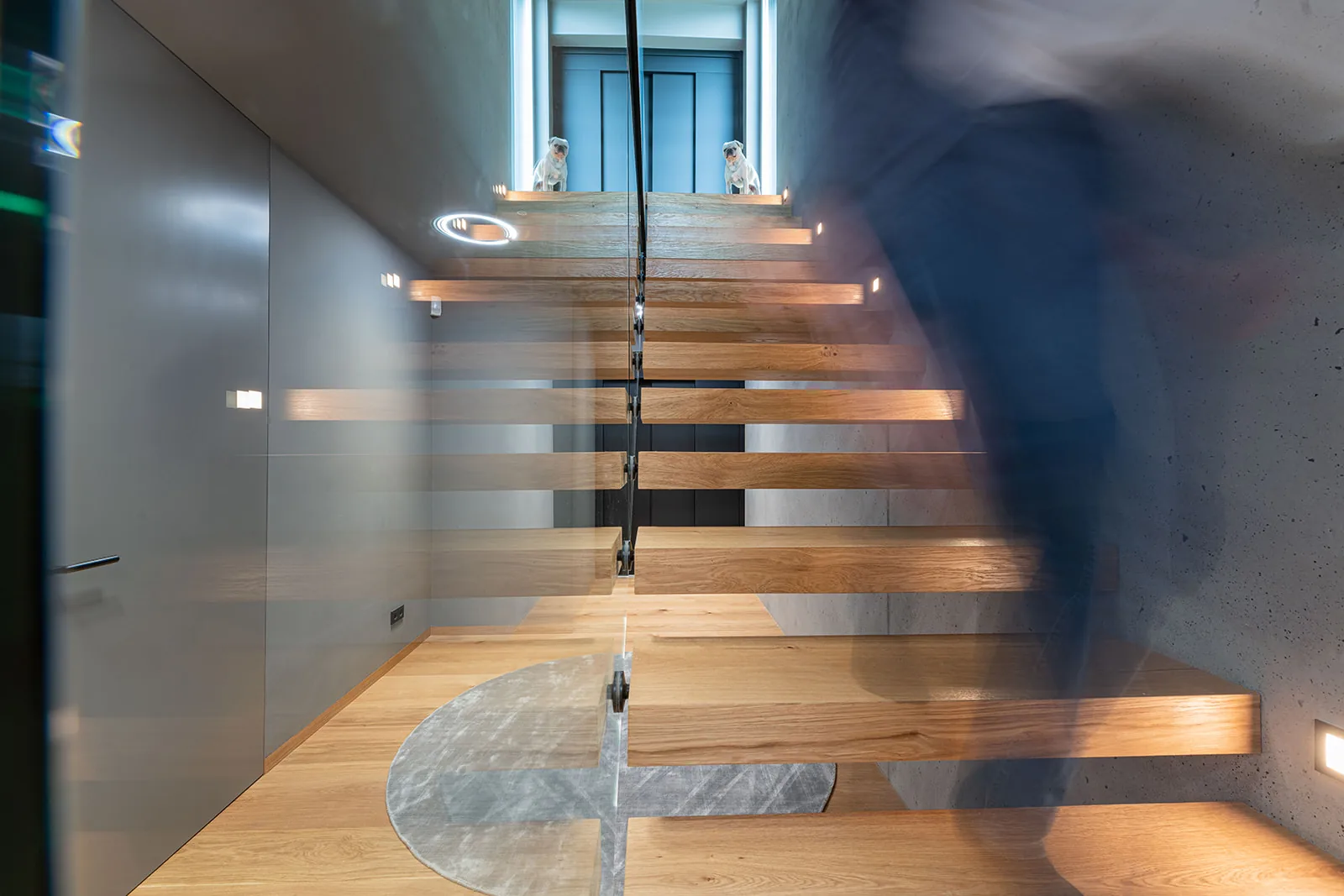 Kragarmtreppe aus Eiche an einer Sichtbetonwand, Ganzglasgeländer mit schwarzem Handlauf; rechts steigt eine Person als Bewegungsunschärfe hinauf, im Eichenboden ein runder Marmoreinsatz.
