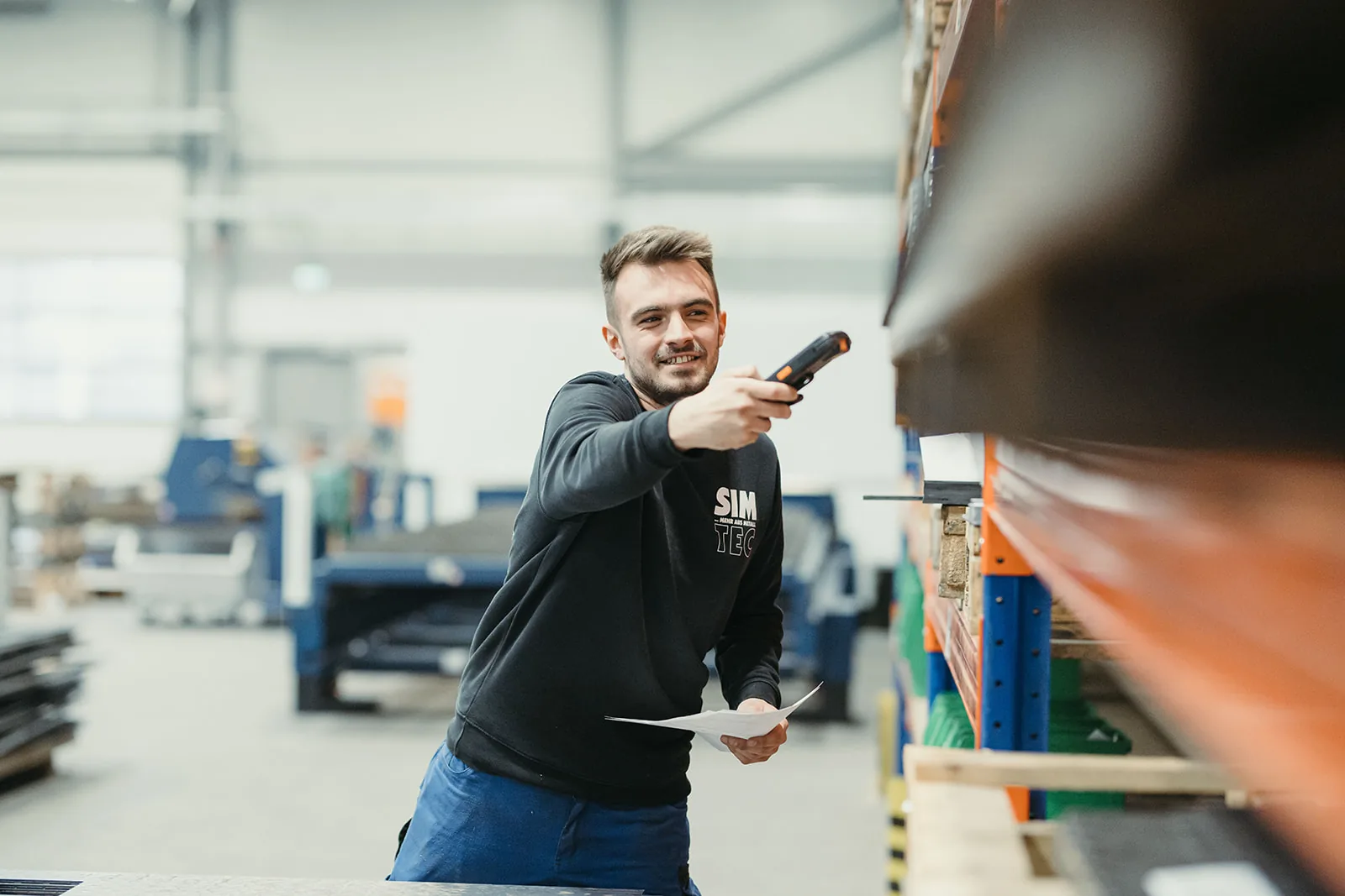 Lagermitarbeiter im Firmen-Hoodie scannt mit einem Handgerät Ware im Regal, im Hintergrund die weite, hell ausgeleuchtete Fertigungshalle.