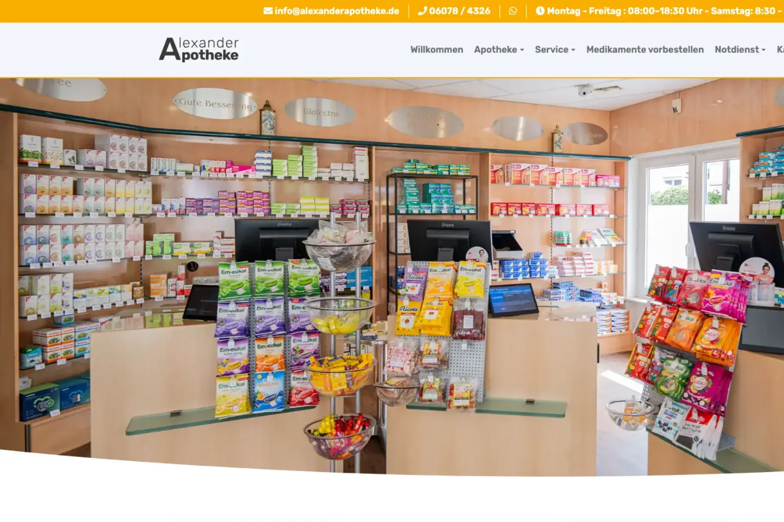 Startseite der Alexander Apotheke: über dem Menü eine orange Kontaktleiste, darunter der Blick in die Offizin mit hölzernen Sichtwahl-Regalen.